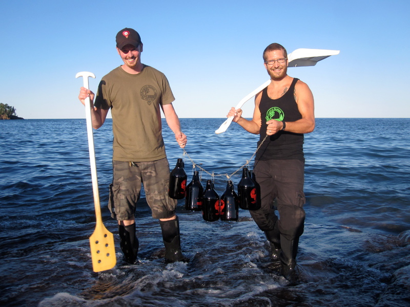 Two men holding a stringer of growlers a paddle, and a shovel in front of Lake Superior.